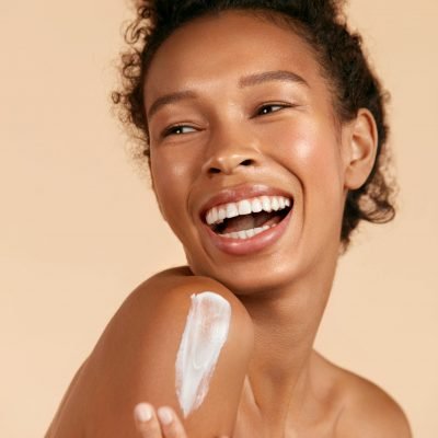 Body skin care. Smiling black woman applying body cream portrait. Beautiful happy african american girl model with cosmetic moisturizing lotion on perfect hydrated soft skin on shoulder at studio